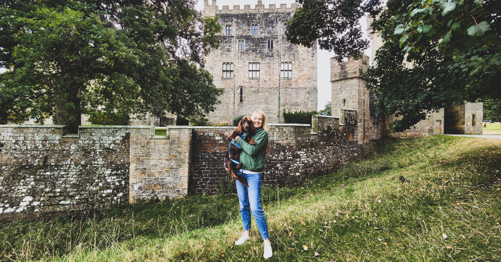 Woman stodd smiling at camera and holding a brown dog with Raby Castle visible in the background.